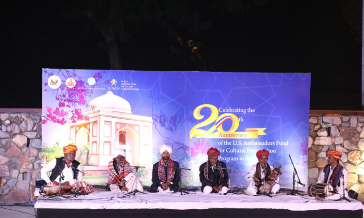 Performers of Indian folk music sitting on stage with a banner mentioning the 20th anniversary of U.S. AFCP projects in India behind them